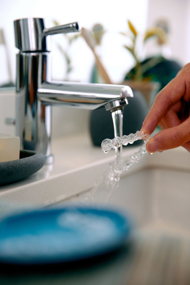 Hands rinsing an Invisalign clear aligner under tap water — proper aligner care and maintenance