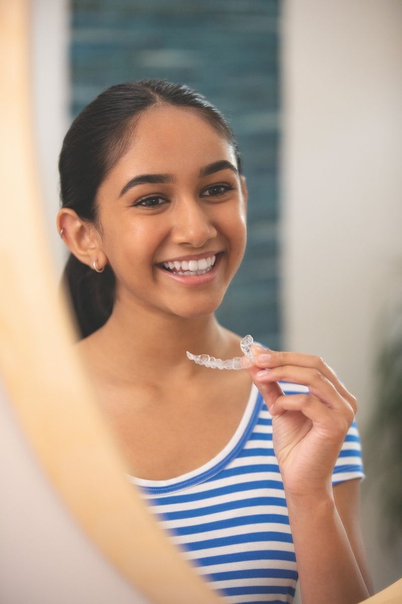Teen girl looking in mirror while inserting her Invisalign aligner — attachments are small and barely visible